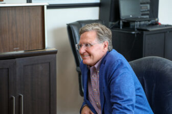 A man in a blue blazer and checkered shirt sits on a chair in an office, smiling and looking to the side. A wooden cabinet and office equipment are in the background.
