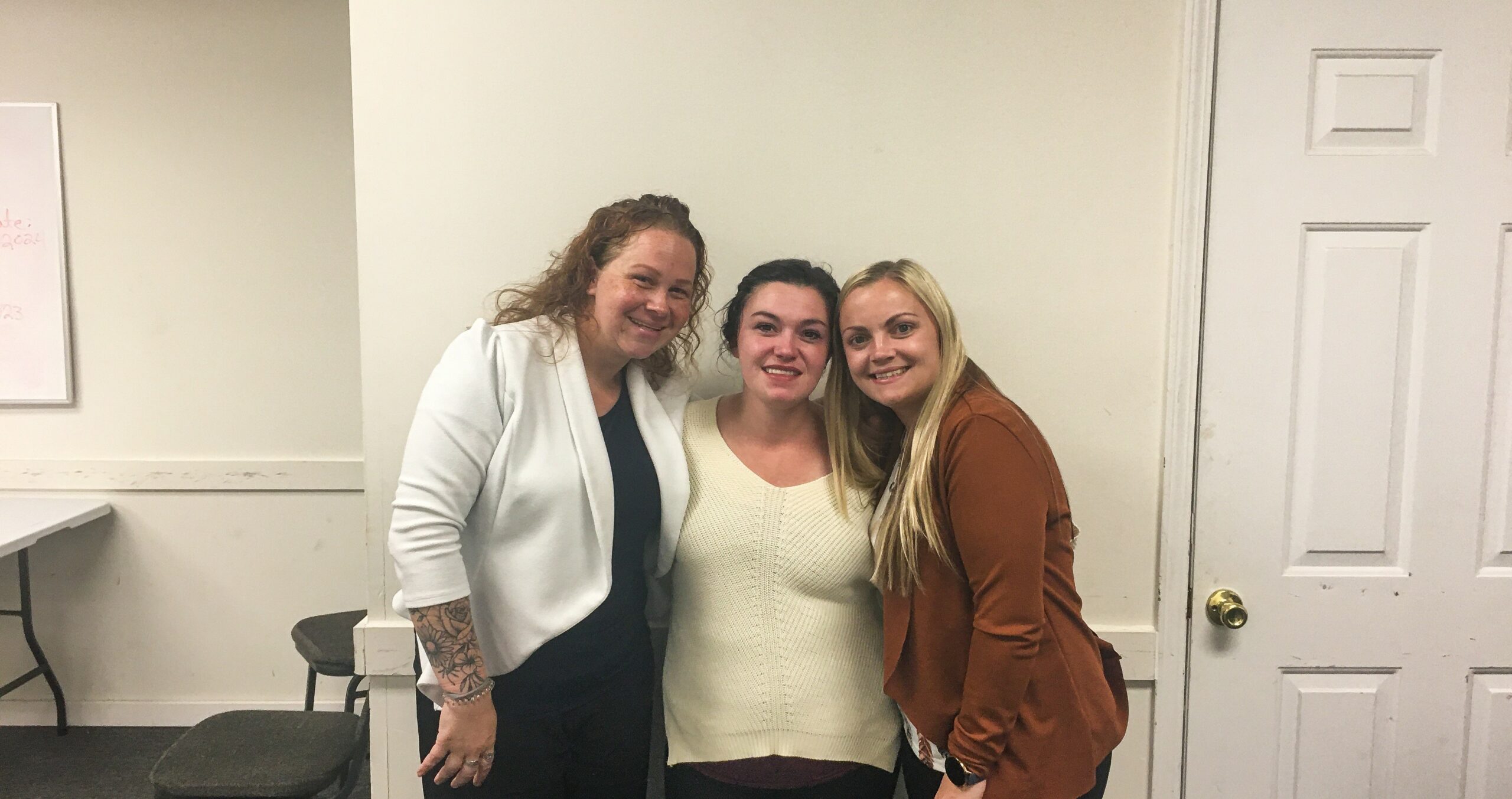 Three women smiling and posing together in a room stand close with arms around each other’s shoulders. A whiteboard is partially visible on the left, and a closed door is on the right.