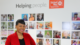 Michelle Tucker, in a red jacket, smiles in front of a wall adorned with the United Way logo and an array of photos. The display highlights the slogan "Helping people," emphasizing the community spirit and volunteer opportunities fostered by this nonprofit organization.