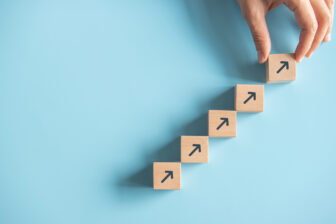 A hand places a wooden block with an upward arrow at the top of a stair-like formation made of similar blocks on a light blue background, symbolizing growth and progress.