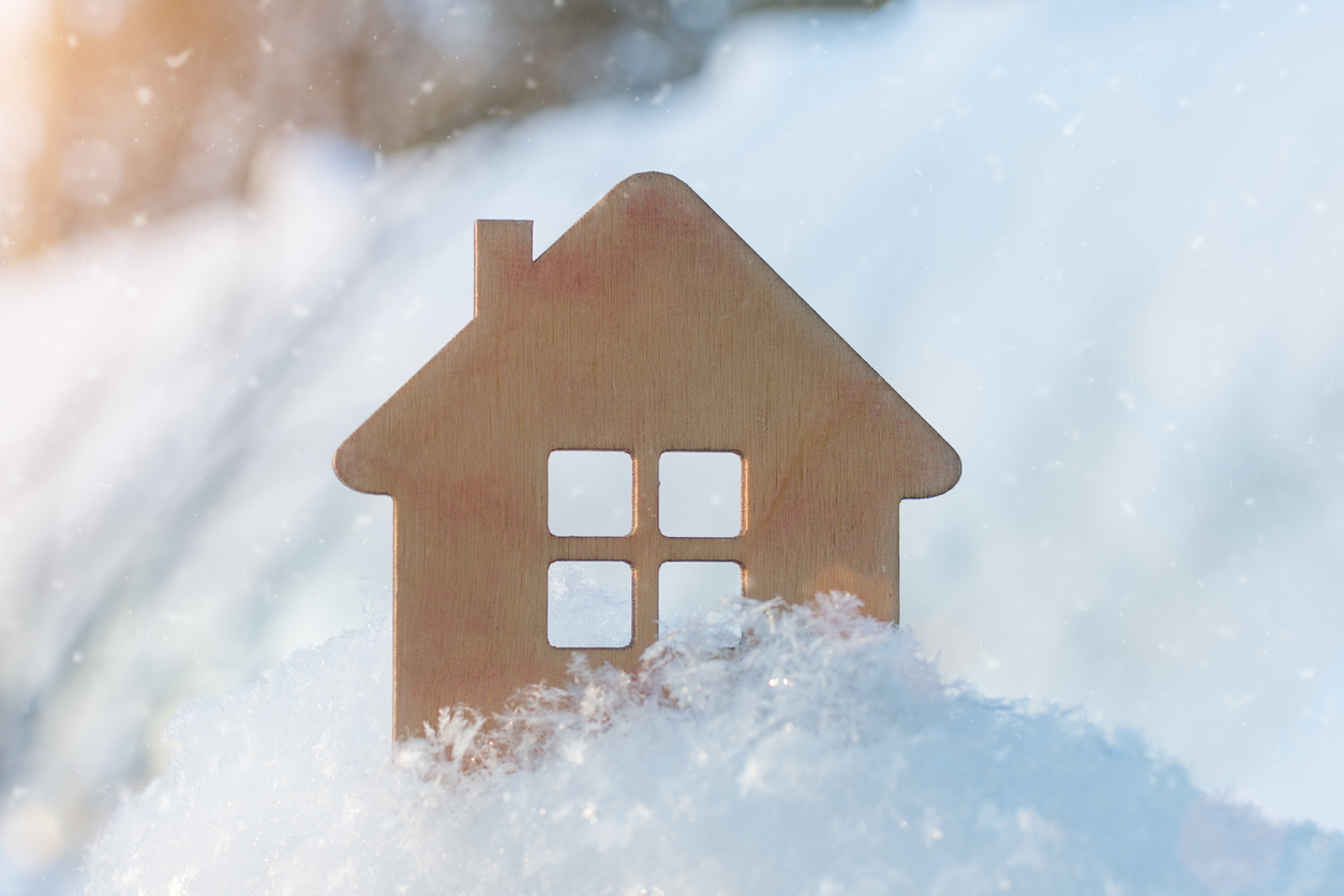 Amidst a snowy landscape, a wooden cutout of a house with windows stands surrounded by gently falling snowflakes. The sun shines softly in the background, casting a warm glow on this cozy winter scene. A sense of community is reflected in the serene beauty of nature's canvas.