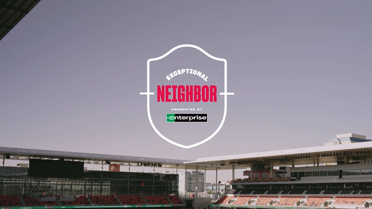 A stadium with empty seats under a clear sky features the words "Exceptional Neighbor presented by Enterprise" prominently in a shield-shaped logo at the center of the image. This scene embodies community spirit and highlights volunteer opportunities, welcoming those ready to make a difference.