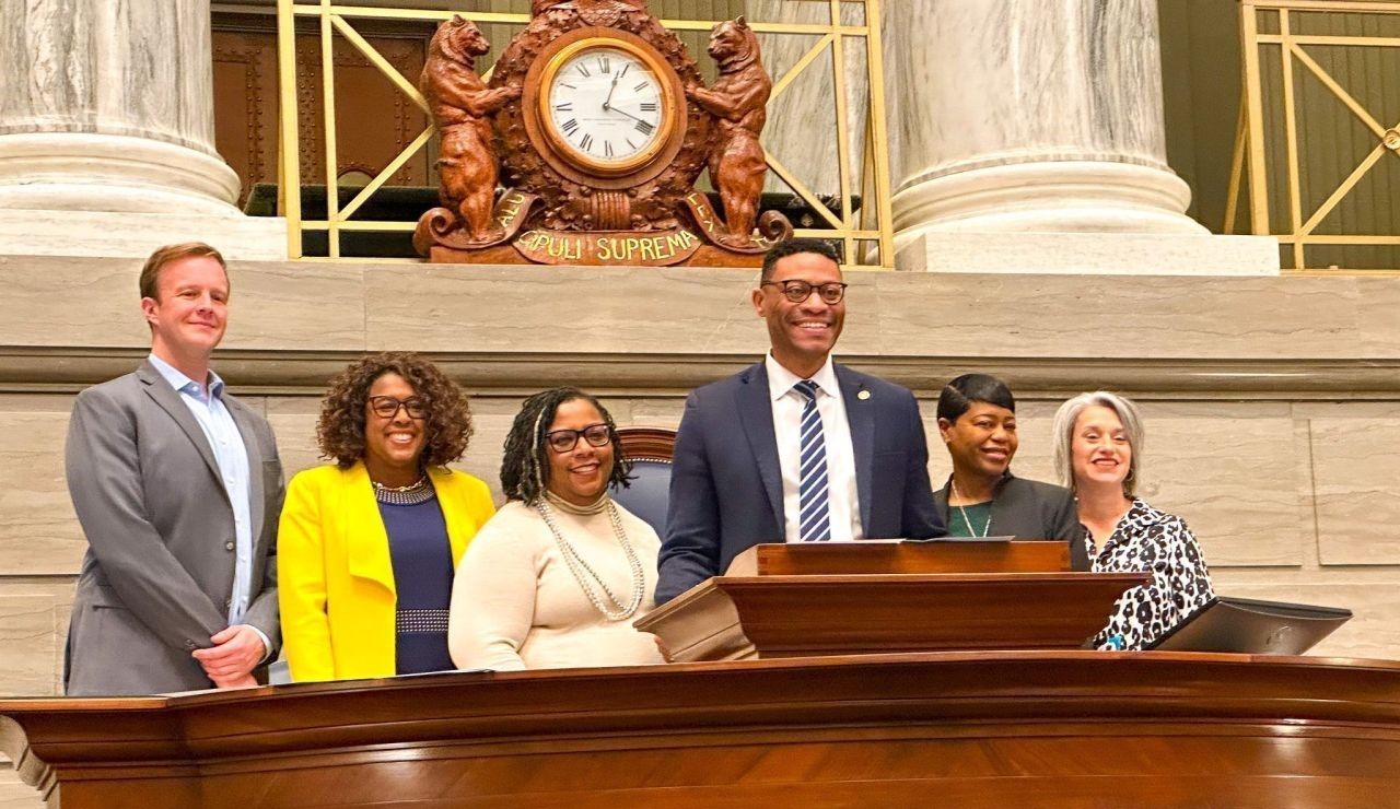 A group of six people, embodying a sense of community, stands together behind a podium in a formal setting. There is a large clock and ornate decorations in the background. The individuals are smiling and dressed in business attire, reflecting camaraderie and professionalism.